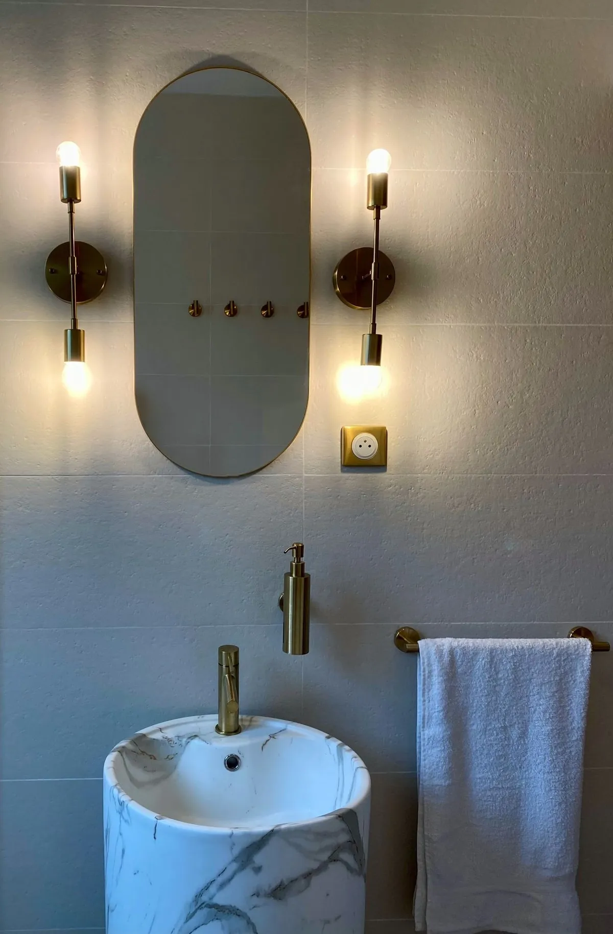 Vasque colonne effet marbre sous un miroir ovale encadré de deux appliques dorées