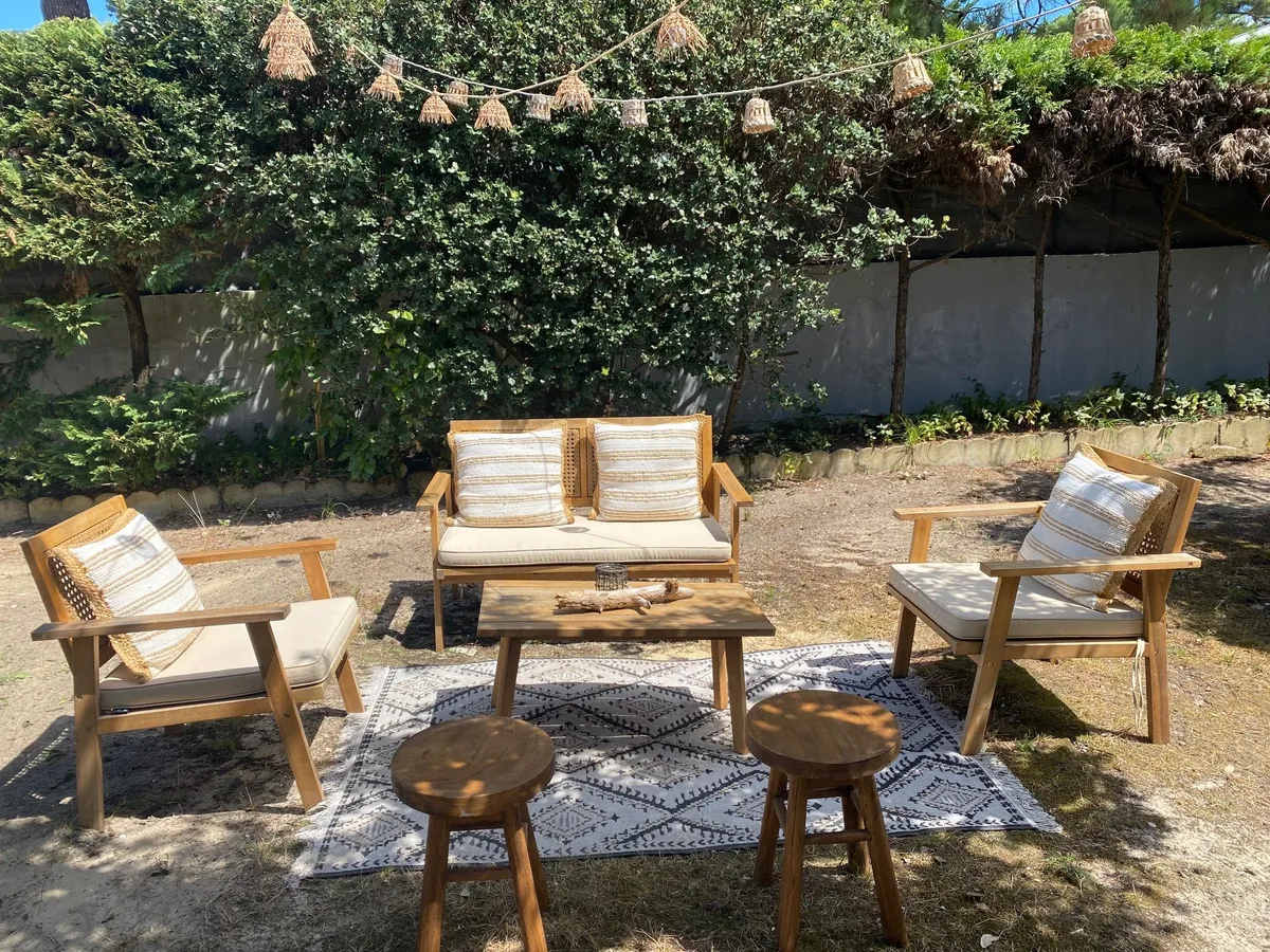Salon de jardin en bois aux coussins écrus sur un tapis d'extérieur, sous une guirlande