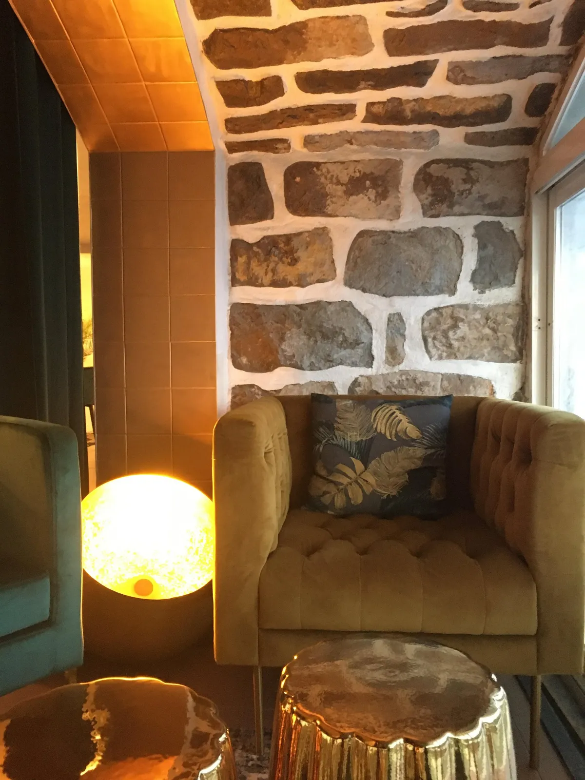 Fauteuil capitonné moutarde contre un mur de pierre, lampe globe et table dorée