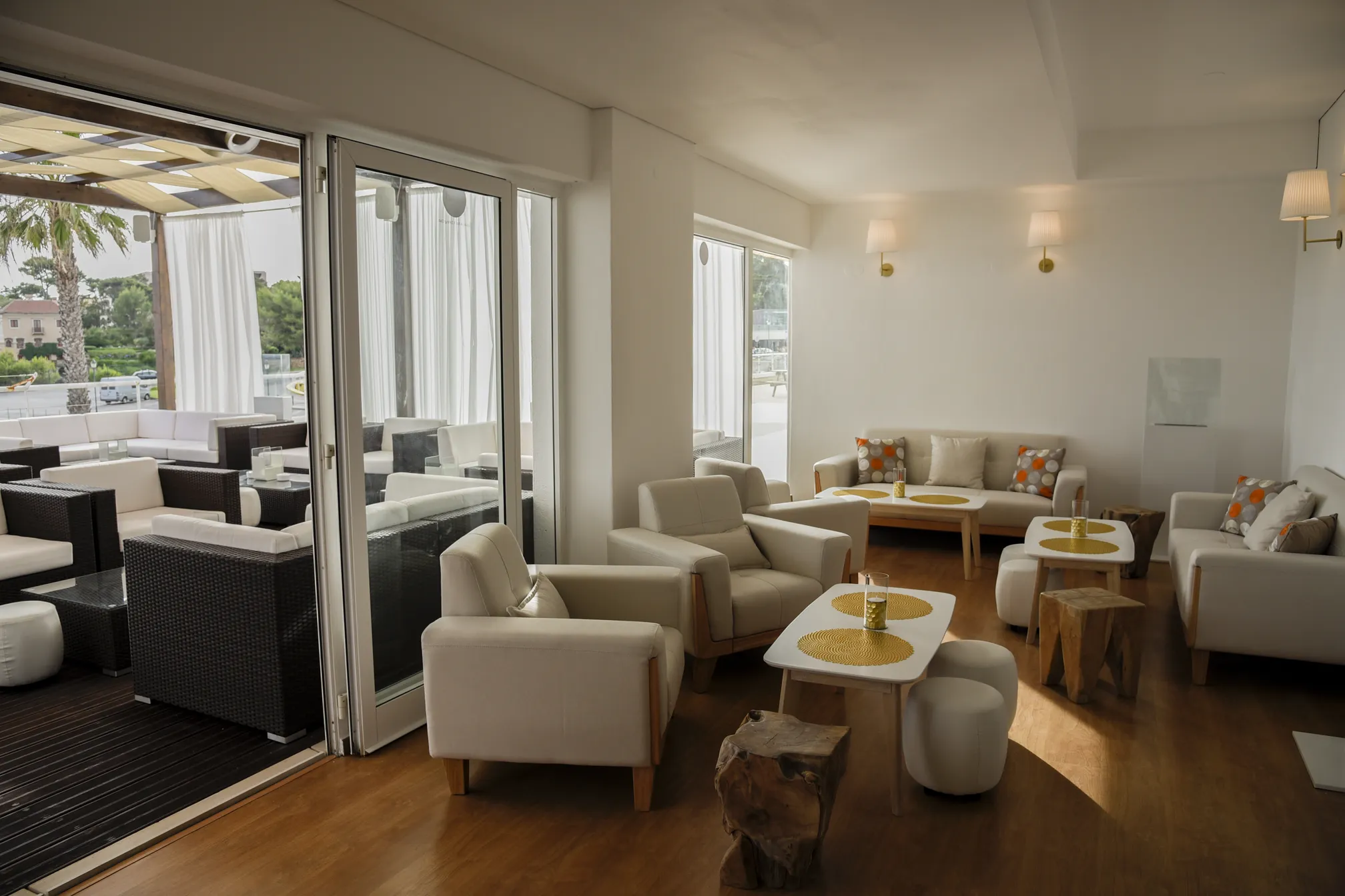 Lounge lumineux aux murs blancs, fauteuils crème et tables basses en bois, ouvert sur une terrasse sous pergola